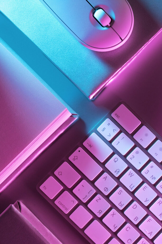 ピンクと水色のネオンに照らされたデスク。キーボード、マウス、ノートが置かれている